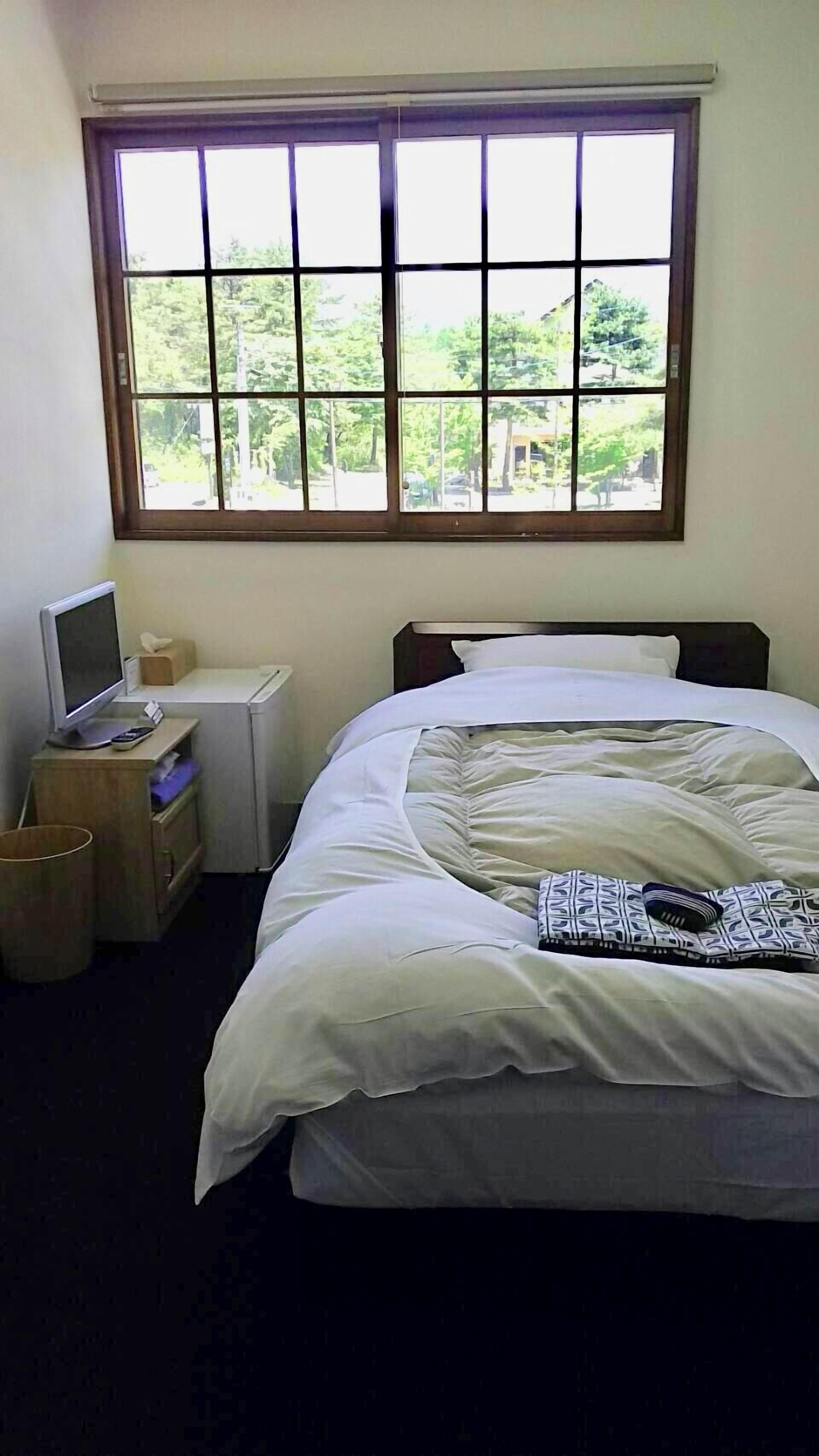 Ein Schlafzimmer mit einem Bett, einem Schreibtisch mit Computer, einem Fenster mit Blick ins Grüne und einer Mülltonne.