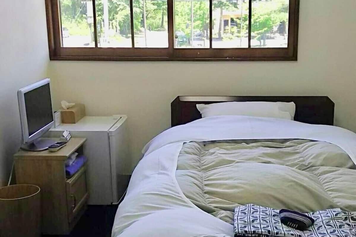 Een slaapkamer met een bed, een bureau met een computer, een raam met uitzicht op groen, en een prullenbak.