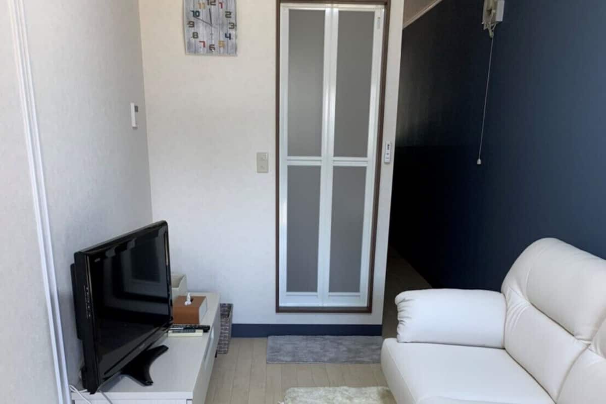 Een woonkamer met een witte bank, een televisie, een klein tafeltje, een witte prullenbak, een zacht vloerkleed en een glazen schuifdeur.