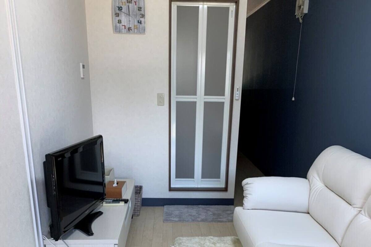 Ein Wohnzimmer mit einem weißen Sofa, einem Fernseher, einem kleinen Tisch, einer weißen Mülleimer, einem flauschigen Teppich und einer Glastür, die sich öffnen lässt.