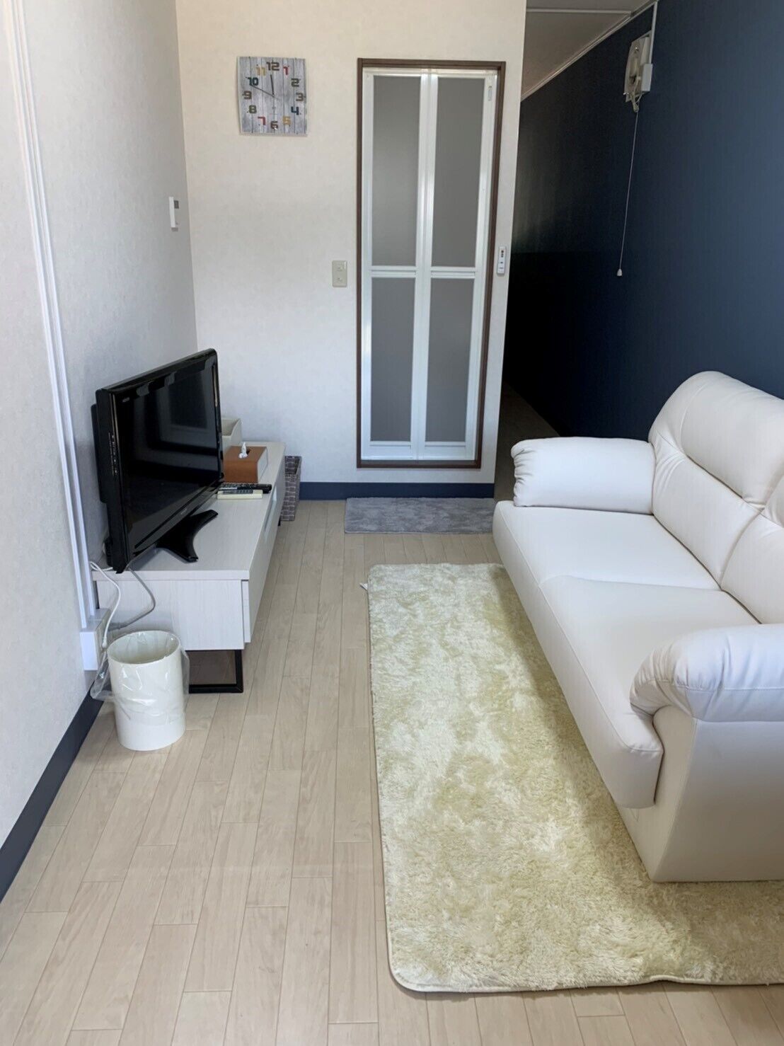 Una sala de estar con un sofá blanco, televisor, una mesita, alfombra y puerta corrediza de vidrio.