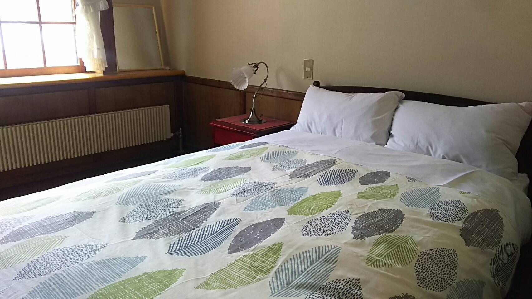 Una cama con una colcha estampada con hojas, una mesita de noche con una lámpara y una ventana con cortinas.