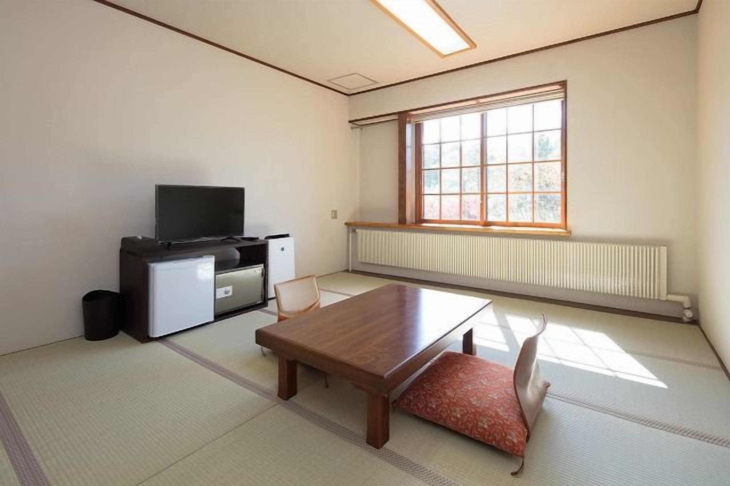 Una habitación con suelo de tatami, una mesa de madera, un televisor sobre un mueble y una ventana con vistas a árboles.