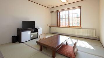 Una habitación con suelo de tatami, una mesa de madera, un televisor sobre un mueble y una ventana con vistas a árboles.