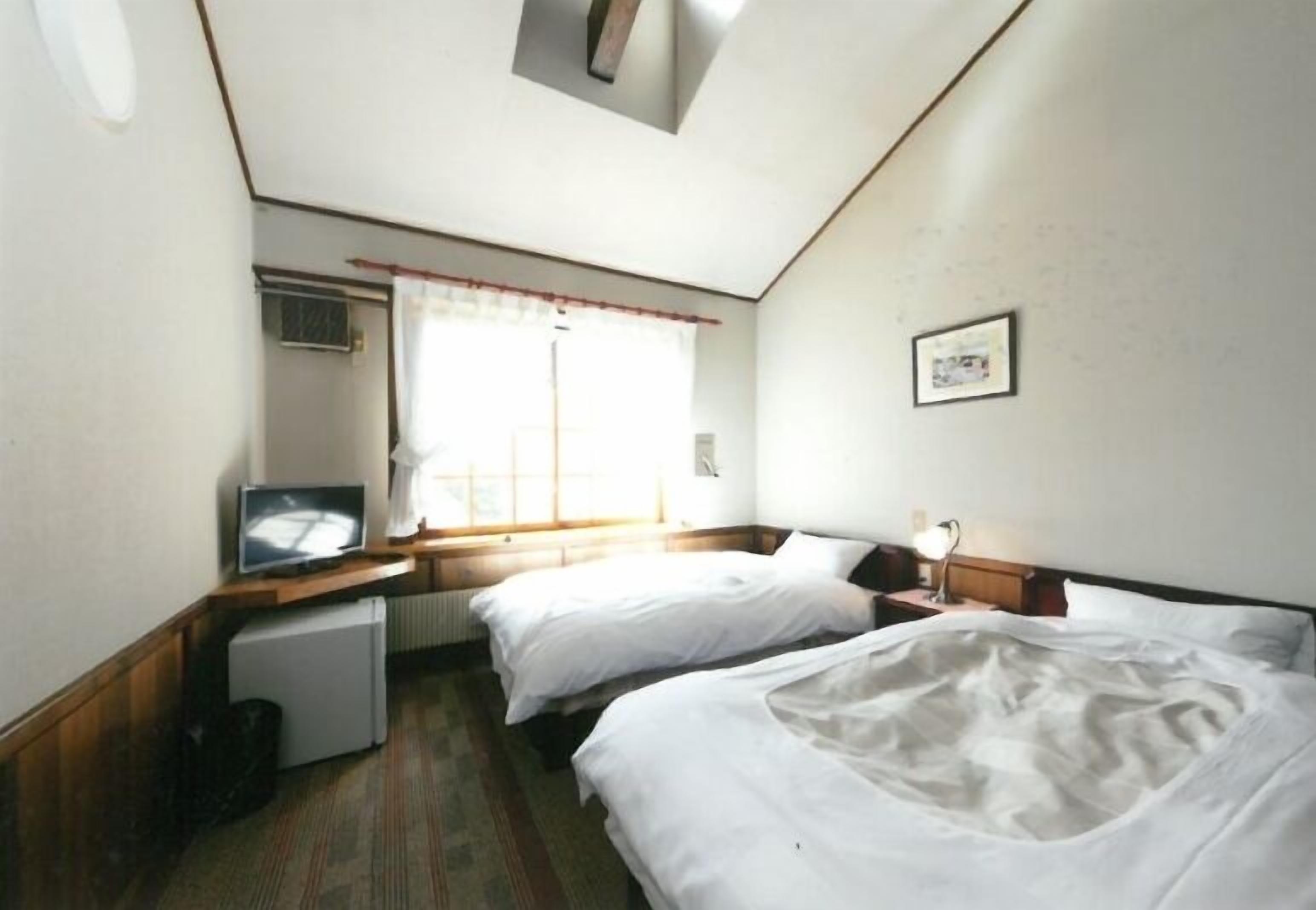 Ein Hotelzimmer mit zwei Betten, einem kleinen Schreibtisch mit Fernseher, einem Heizkörper und einem Fenster mit Vorhängen.