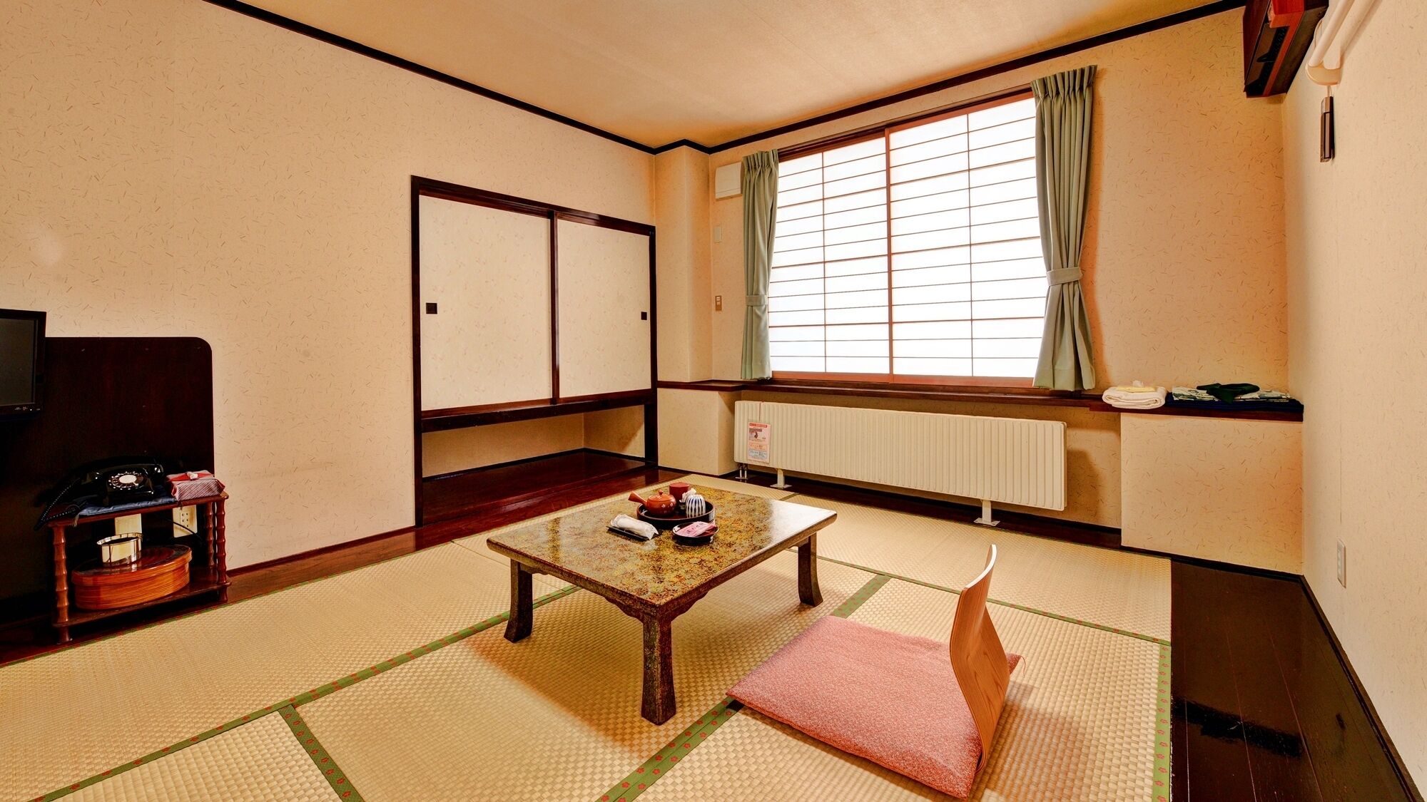 Una habitación japonesa tradicional con suelo de tatami, una mesa de madera baja, una cama futón, un radiador y una ventana con cortinas.