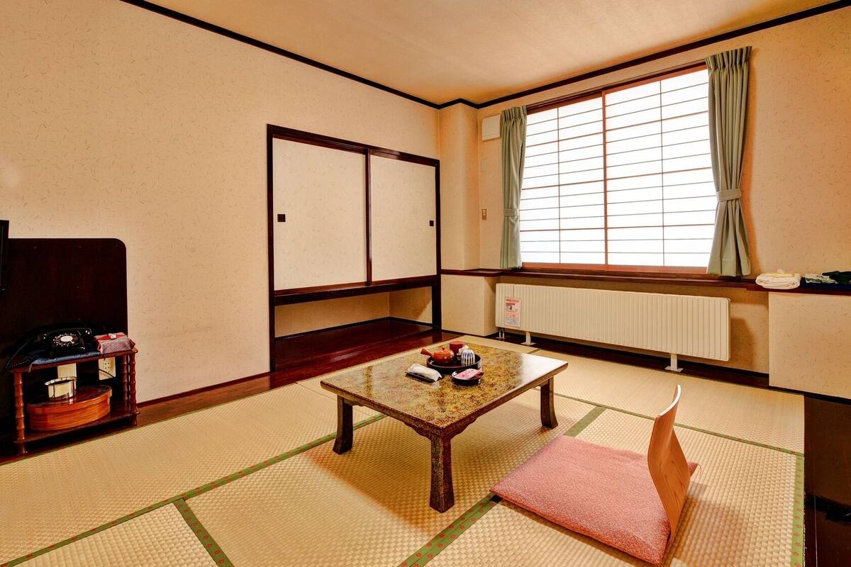 Una habitación japonesa tradicional con suelo de tatami, una mesa de madera baja, una cama futón, un radiador y una ventana con cortinas.