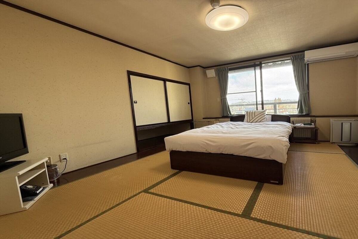 Habitación de hotel con cama, televisor, ventana con cortinas, radiador y vista a la ciudad.
