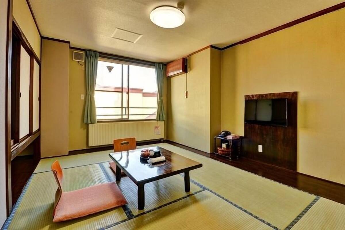 Una habitación con suelo de tatami, una mesa baja con cojín, una silla, un televisor montado en la pared y una ventana con cortinas.