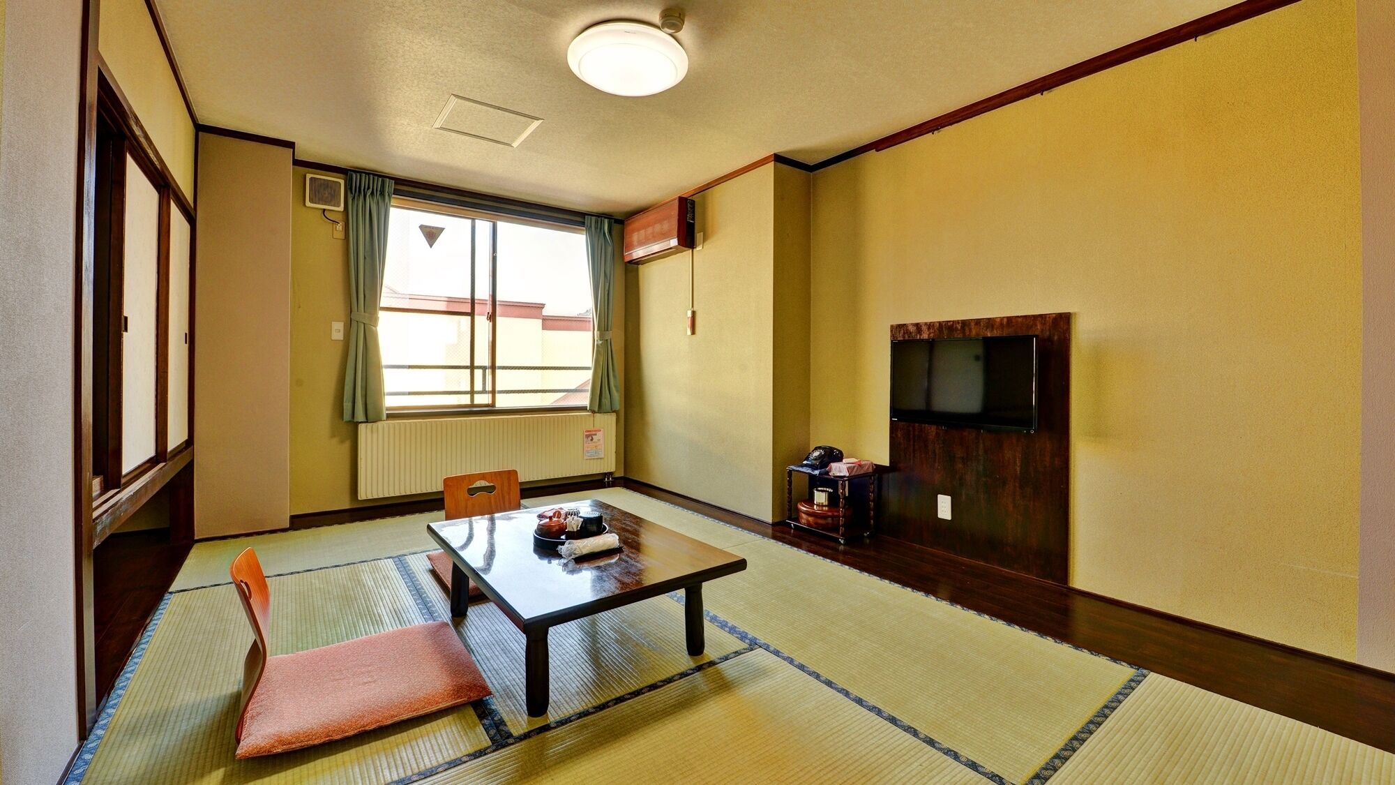 Una habitación japonesa tradicional con suelo de tatami, una mesa baja, una silla, un televisor montado en la pared y una ventana con cortinas.