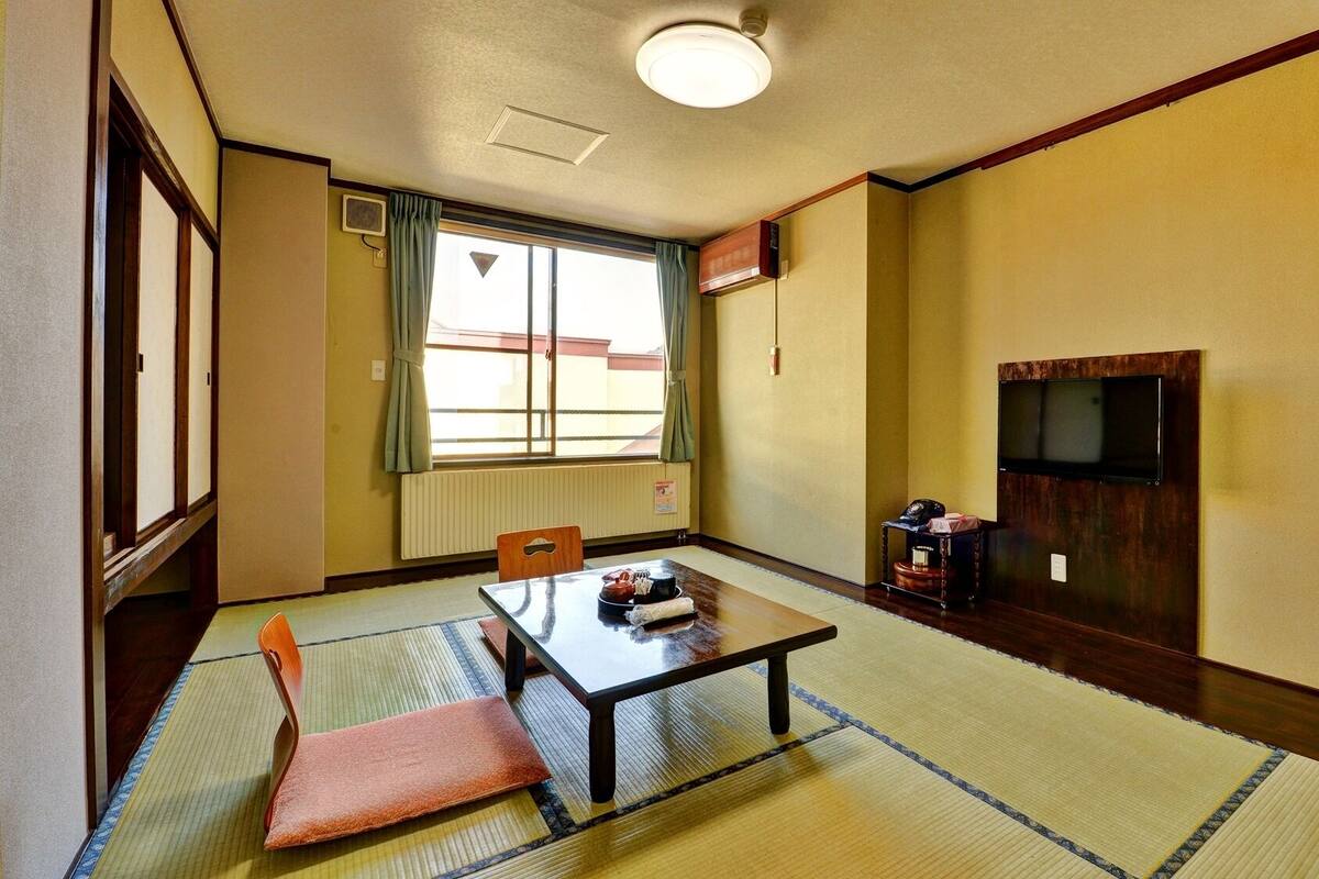Una habitación japonesa tradicional con suelo de tatami, una mesa baja, una silla, un televisor montado en la pared y una ventana con cortinas.