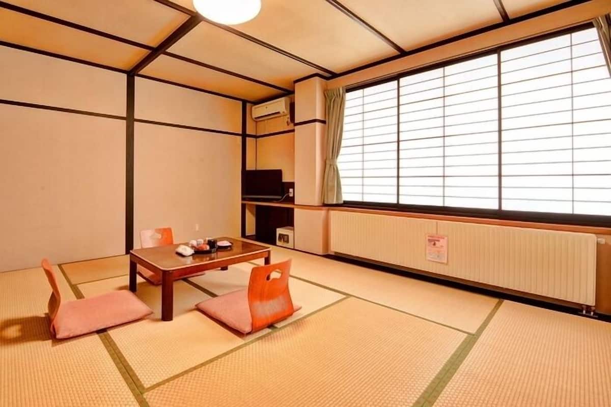 Una habitación japonesa tradicional con suelo de tatami, una mesa baja con cojines, un televisor y una ventana con persianas.