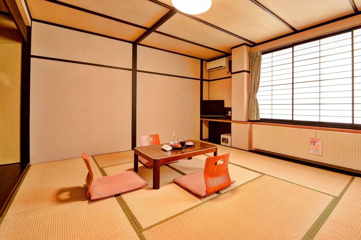 Una habitación japonesa tradicional con suelo de tatami, una mesa baja con cojines, un televisor y un ventanal con paneles shoji.