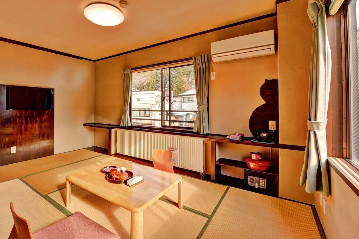 Una habitación tradicional japonesa con suelo de tatami, una mesa de madera baja, un televisor y una ventana con cortinas.