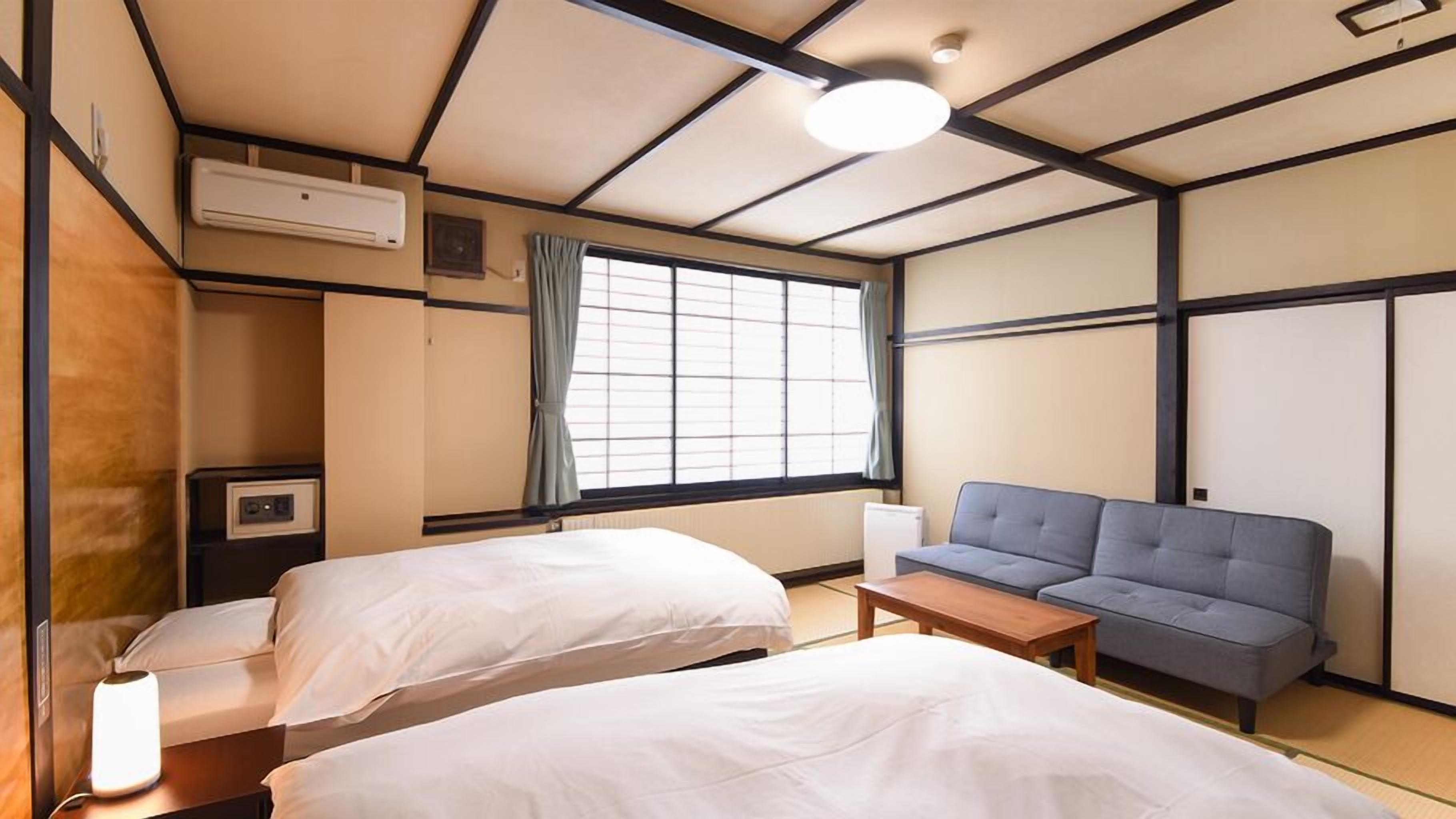 Habitación de hotel con dos camas, un sofá, una mesita, una ventana con cortinas, un equipo de aire acondicionado y un televisor de pared.