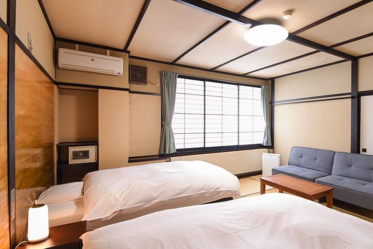 Habitación de hotel con dos camas, un sofá, una mesita, una ventana con cortinas, un equipo de aire acondicionado y un televisor de pared.