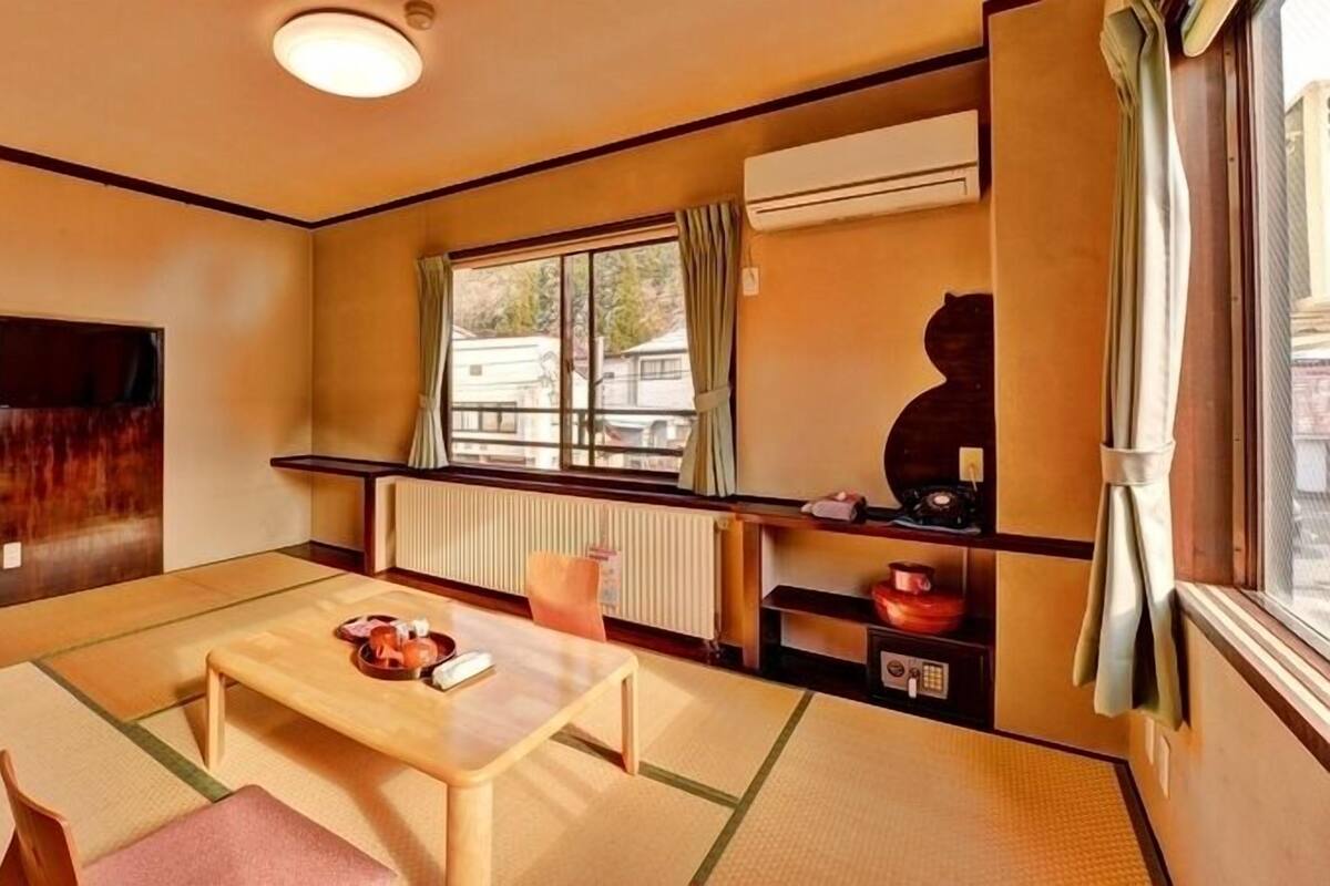 Una habitación tradicional japonesa con suelo de tatami, una mesa baja, un futón como cama, una ventana con cortinas y un radiador.