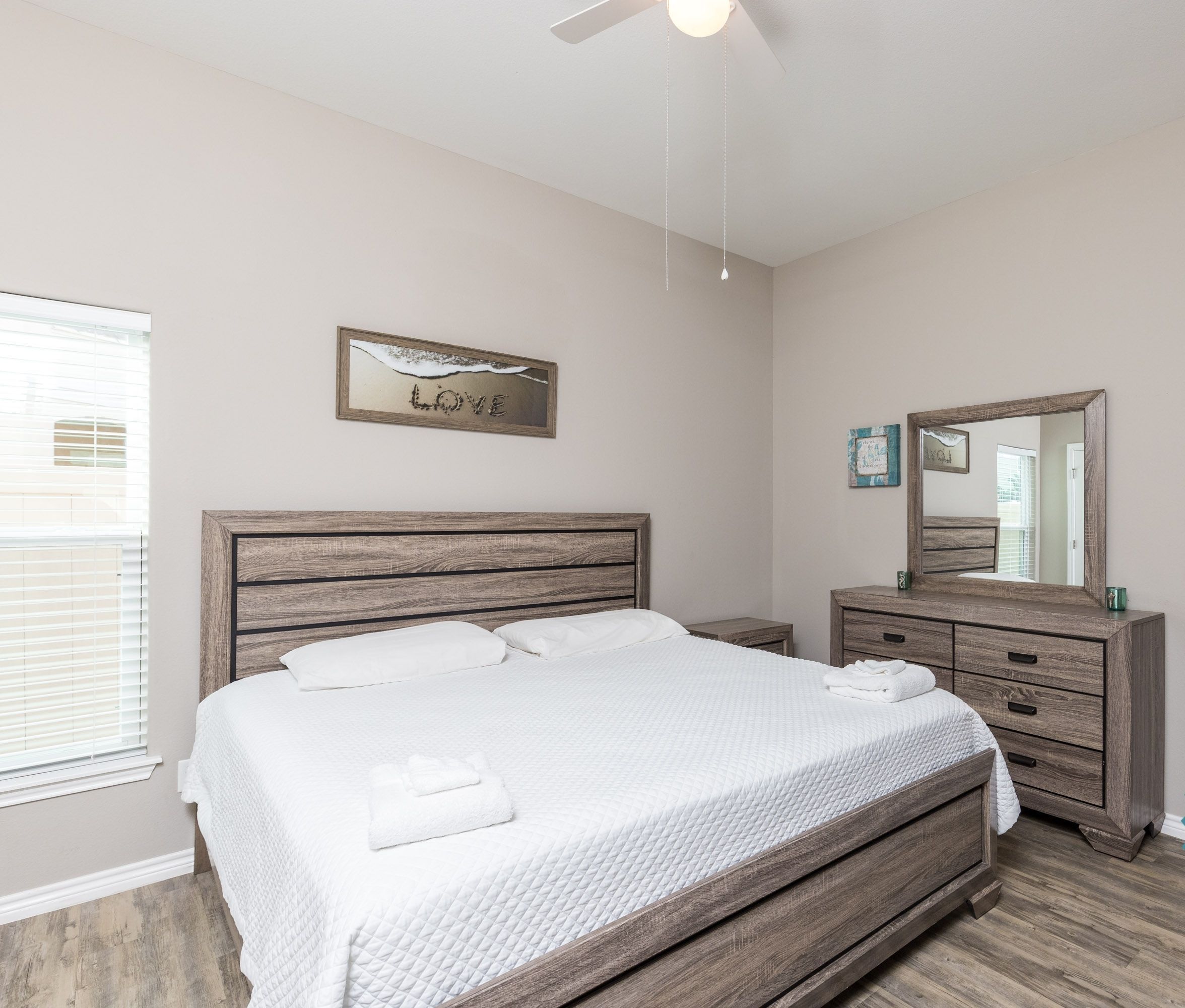 A clean, modern bedroom with a wooden bed, a nightstand, a dresser, and a ceiling fan.
