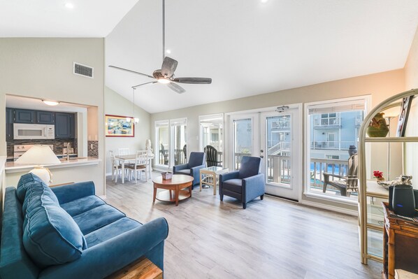 A spacious living area with a blue sofa, a dining table, and chairs, a ceiling fan, and large windows offering a view of the outside.