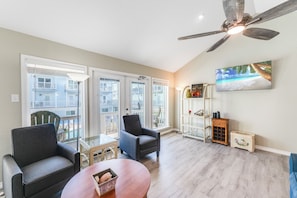 Un salon spacieux doté d’un ventilateur de plafond, d’une grande fenêtre offrant une vue sur un immeuble et d’un tableau représentant une scène de plage.