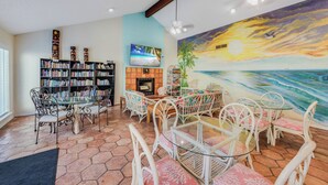 A living room with a fireplace, a large bookshelf, a dining area with white chairs, and a mural of a sunset over the ocean. - Beachside Condo | Pool, Hot Tub & Private Balconies (Corpus Christi)