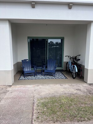 Ein überdachter Patio-Bereich mit drei blauen Adirondack-Stühlen, einem gemusterten Teppich und einem Fahrrad, das an der Wand steht.