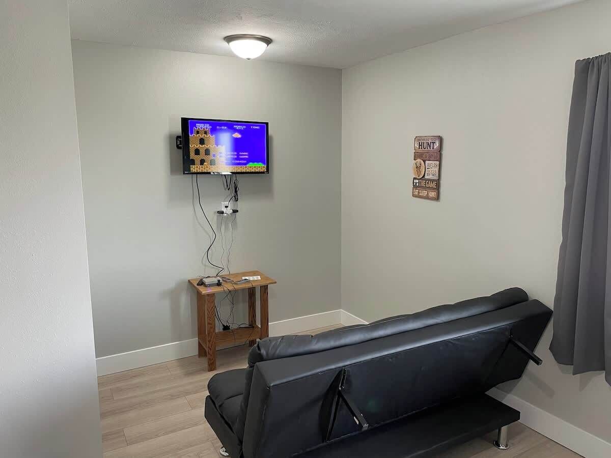 A room with a black leather couch, a television mounted on the wall, a small wooden side table, and a sign on the wall.