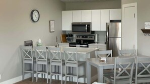 Een kleine keuken met witte kastjes, een magnetron, een koelkast en een klok aan de muur.