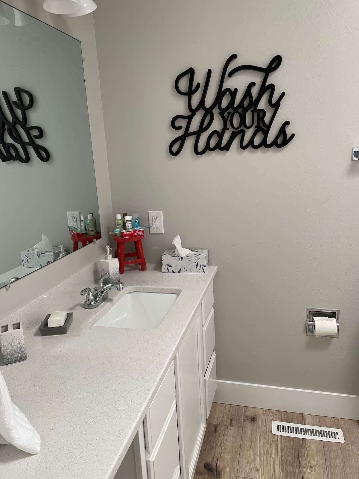 Un baño con lavamanos doble, un espejo, un letrero que dice 'Lávate las manos', un taburete rojo y un dispensador de papel higiénico.