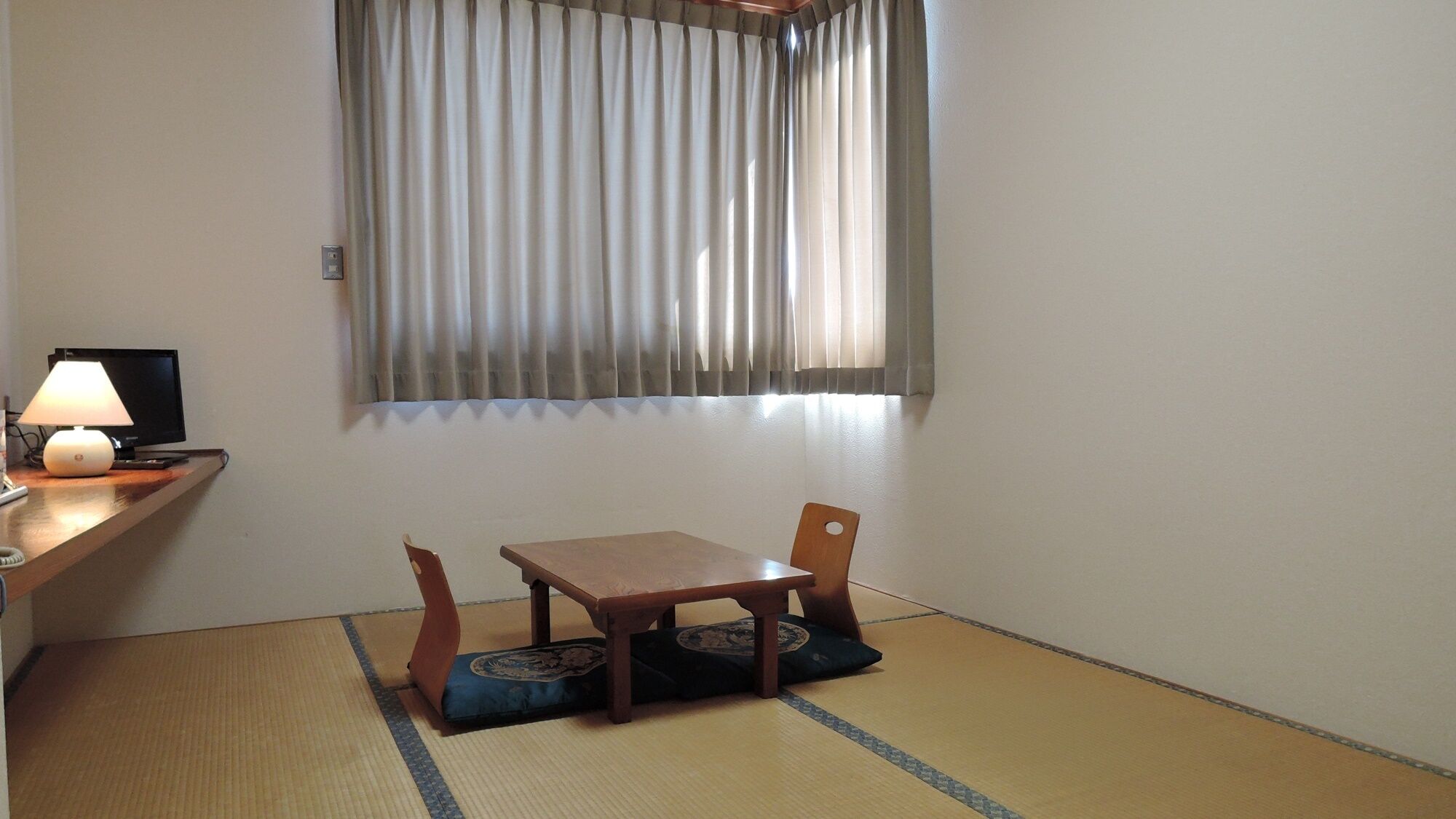 Una habitación con suelo de tatami, una mesa de madera con cojín, una silla, una lámpara y un televisor sobre un soporte.