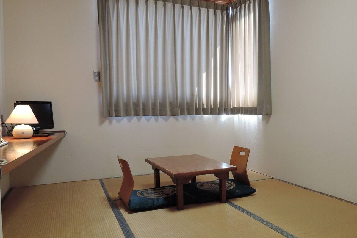 Une pièce avec un sol recouvert de tatamis, une table en bois avec un coussin, une chaise, une lampe et un téléviseur posé sur un meuble.
