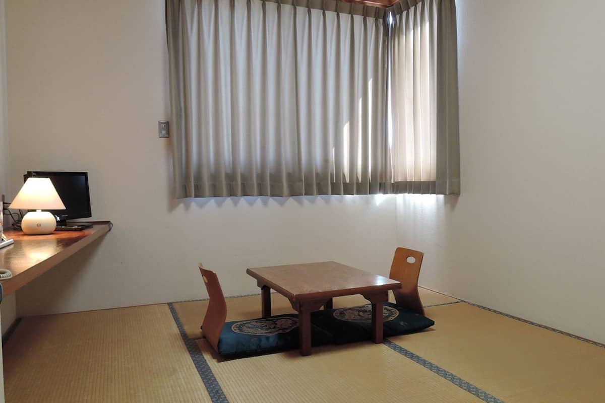 Una habitación con suelo de tatami, una mesa de madera con cojín, una silla, una lámpara y un televisor sobre un soporte.