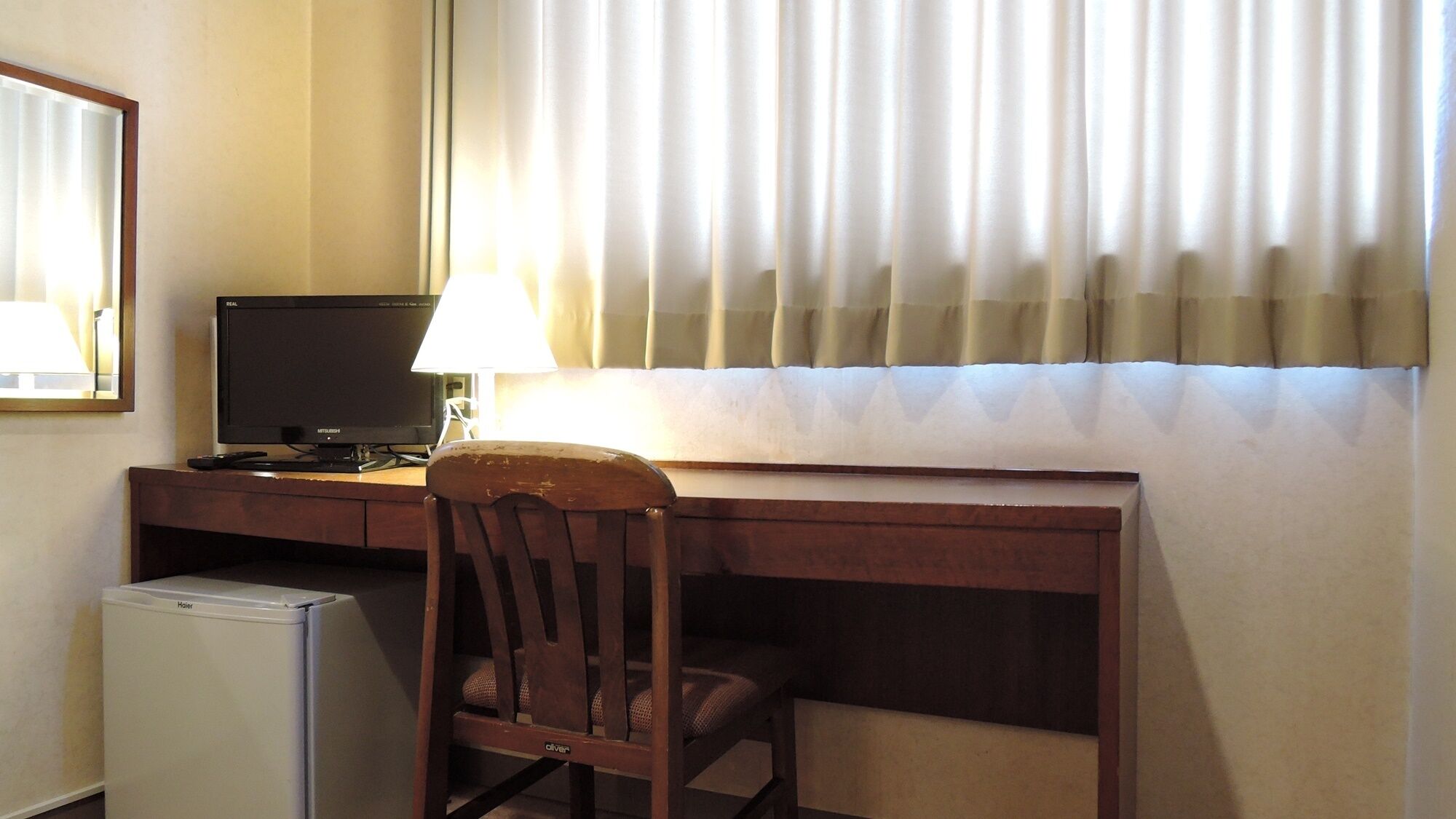 Een hotelkamer met een houten bureau, een televisie, een kleine koelkast, een stoel en een raam met gordijnen.