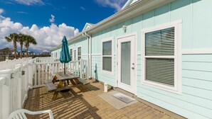 Un bâtiment d’un bleu clair avec des volets blancs, un patio couvert avec une table et des chaises, et une clôture blanche.