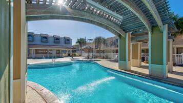 Ein Swimmingpool mit einer Holzpergola darüber, umgeben von Gebäuden und wolkenlosem Himmel.