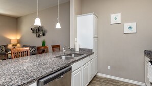 A modern kitchen with granite countertops, white cabinetry, and a built-in dishwasher. There are two pendant lights, a refrigerator, and a dining area with chairs and a table. - Family Beach Townhome | 2 Pools & Walk to Beach (Corp Christi)