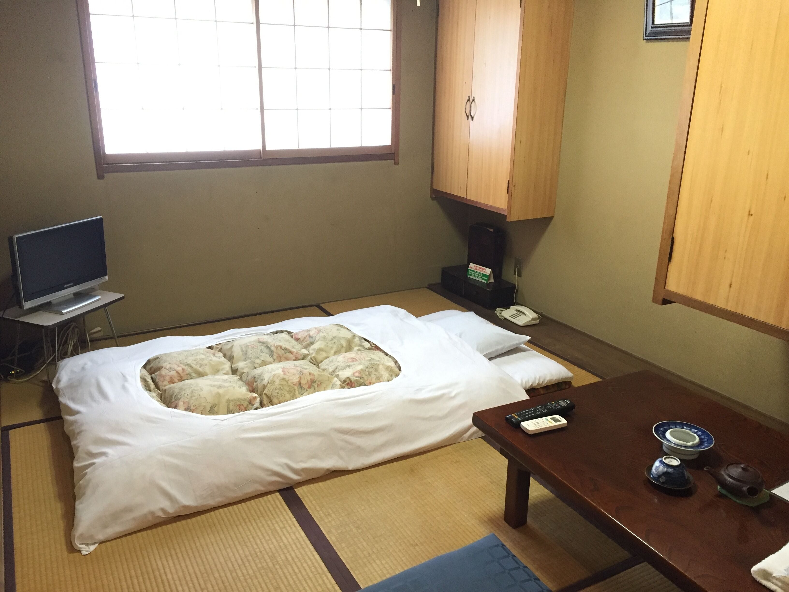 A room with a bed, a television, a wooden cabinet, a telephone, and a table with a teapot and a remote control.