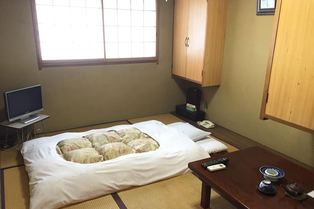 A room with a bed, a television, a wooden cabinet, a telephone, and a table with a teapot and a remote control.