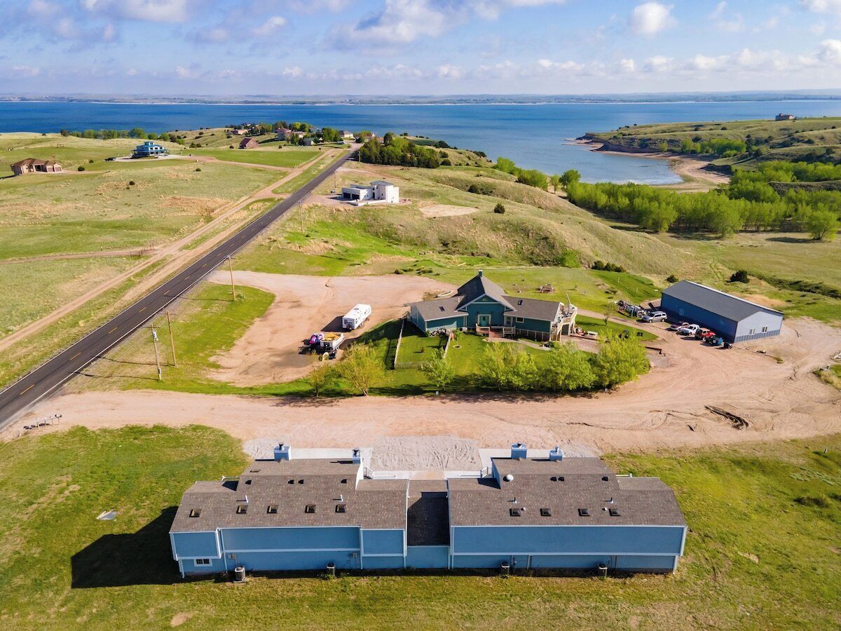 A large industrial building with a flat roof, surrounded by open fields and a road leading to a body of water.
