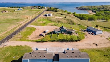 A large industrial building with a flat roof, surrounded by open fields and a road leading to a body of water.