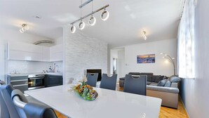 Une cuisine moderne avec des meubles blancs et un four encastré, un coin repas avec une table et des chaises blanches, et un salon avec un canapé et un tableau au mur.