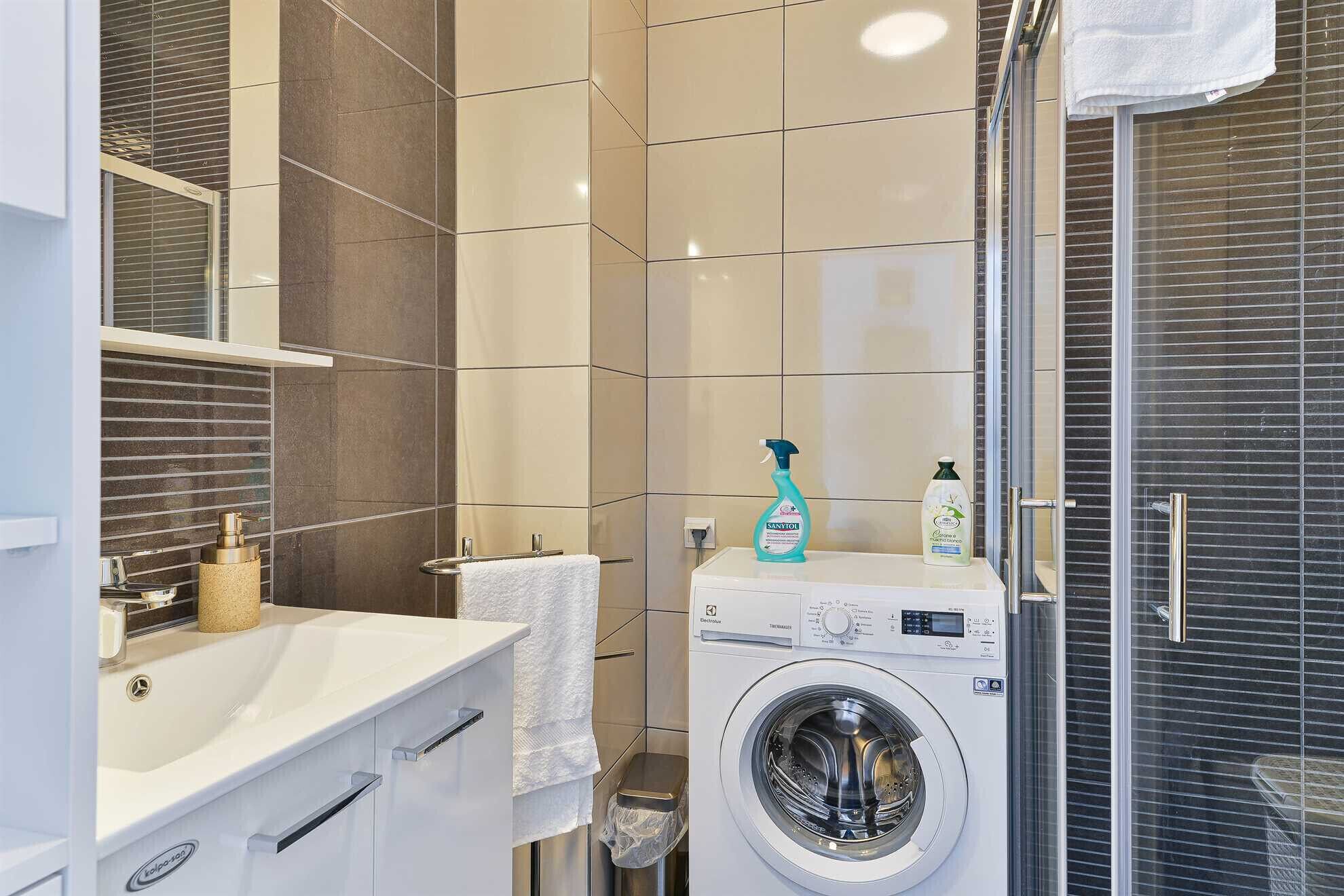 A modern bathroom with a white sink, a washing machine, and a glass shower enclosure.