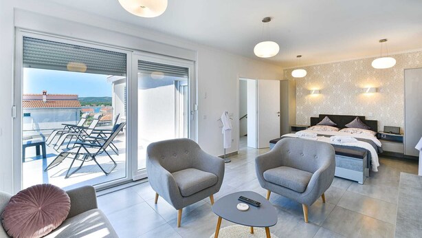Une chambre d’hôtel moderne avec un balcon, deux fauteuils, un lit avec une tête de lit et une petite table avec une télécommande.