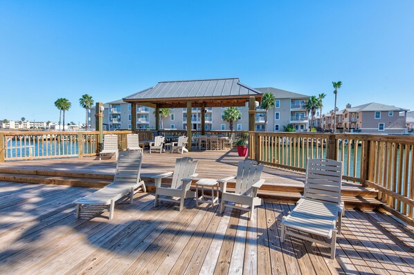 A wooden deck with lounge chairs and a covered seating area, overlooking a body of water and a row of buildings. - Fisherman’s Dream | Waterfront w/ Pool, Hot Tub & Boat Slip (Corpus Christi)