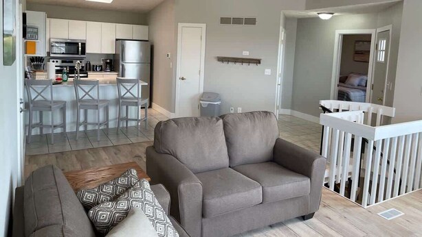 A living room with a grey sofa, a patterned throw pillow, and a wooden floor. There is a kitchen in the background with white cabinets and a microwave. - Bayview Combo Condos 3 & 4, Ogallala Beach (Ogallala)