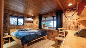 A wooden cabin bedroom with a large bed, a wooden desk, and a window with a view of a staircase.