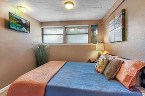 Un dormitorio con cama, televisor montado en la pared, ventana con persianas y un cuadro en la pared.