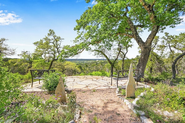 Un chemin de pierres mène à un espace de repos agrémenté de bancs, le tout étant entouré d’arbres et de verdure.