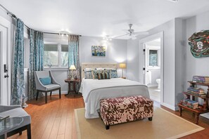 A bedroom with a bed, a chair, a bedside table, a lamp, a ceiling fan, a window with curtains, a painting, a bookshelf, and a bathroom door. - La Casita at Jubilee I 2 BR, stargazing (Wimberley)