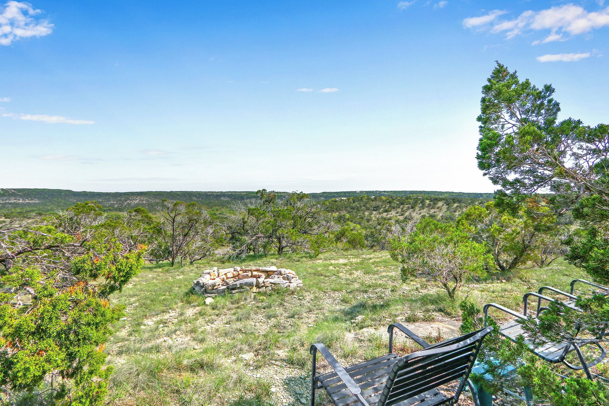 A park with a stone fire pit surrounded by trees, a metal bench, and a clear view of the landscape.